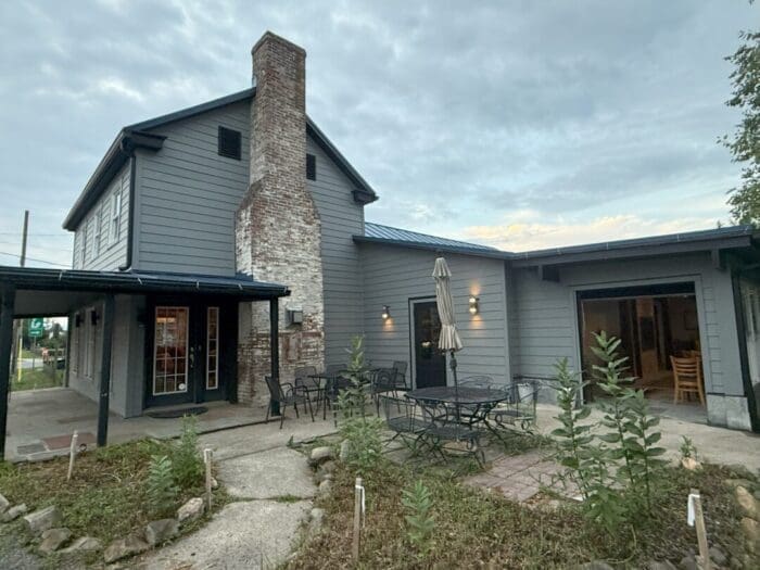 A gray two-story house with a large brick chimney stands under a cloudy sky, resembling the charm of restaurants in Frederick MD. Outdoor tables with umbrellas and garden beds create an inviting patio, while glass doors reveal a warmly lit interior.