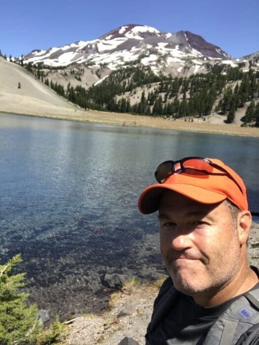 a Man in an Orange Cap and Sunglasses Smiles Near a Clear Blue Alpine Lake Behind Him Evergreen Trees and a Snow dappled Mountain Rise Under a Bright Blue Sky Suggesting a Sunny Day in a Scenic Wilderness Location
