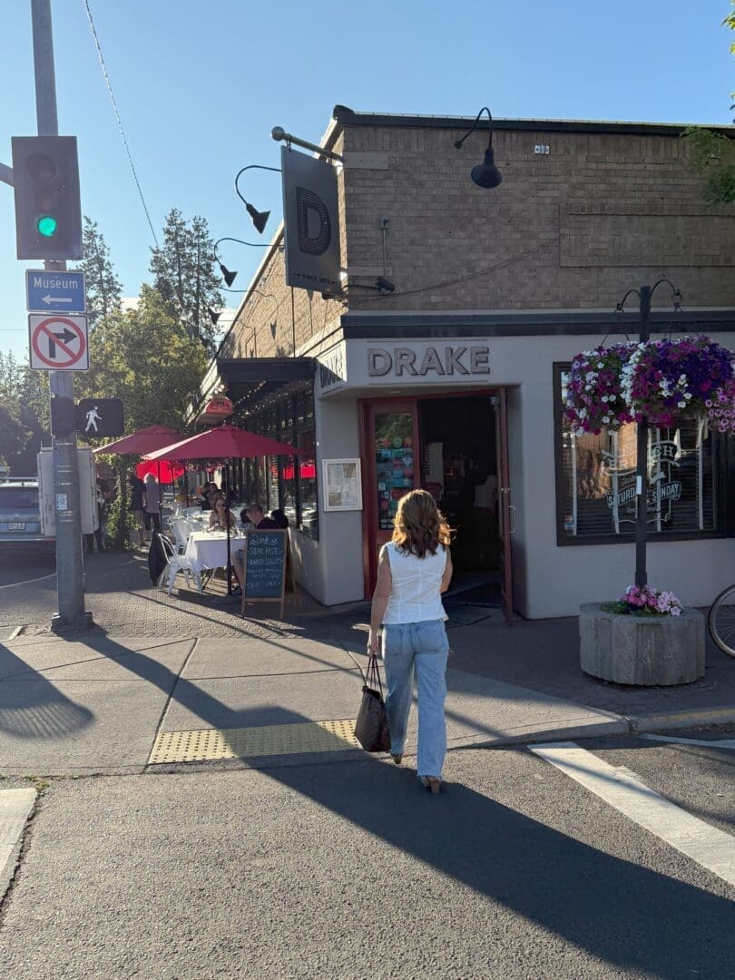 a Woman in a White Top and Light Blue Jeans Walks Toward the Entrance of Drake Restaurant in Downtown Bend Oregon on a Sunny Day Outdoor Diners Sit Under Red Umbrellas As She Explores Airbnb Bend Oregon Nearby Hanging Flower Baskets and a Green Traffic Light Are Visible