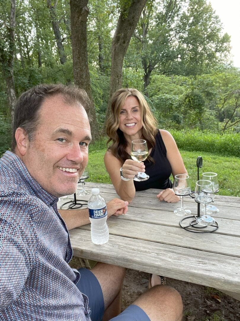 a Man and Woman Sit at a Wooden Picnic Table Outdoors at Market Table Bistro Smiling at the Camera the Woman Holds a Wine Glass with More Empty Glasses on the Table the Man Holds a Water Bottle Amid Lush Green Trees and Grass on a Sunny Day