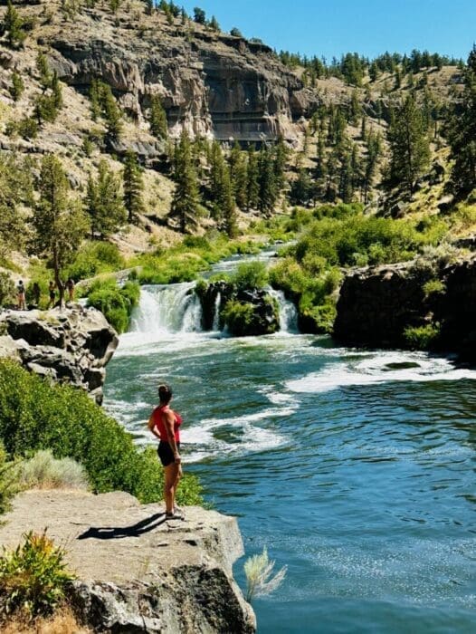 a Person in a Red Shirt and Black Shorts Stands on a Rocky Ledge by Steelhead Falls Gazing at a Small Waterfall Green Trees and Shrubs Surround the Scene with Steep Rocky Hills Under a Clear Blue Sky in the Background