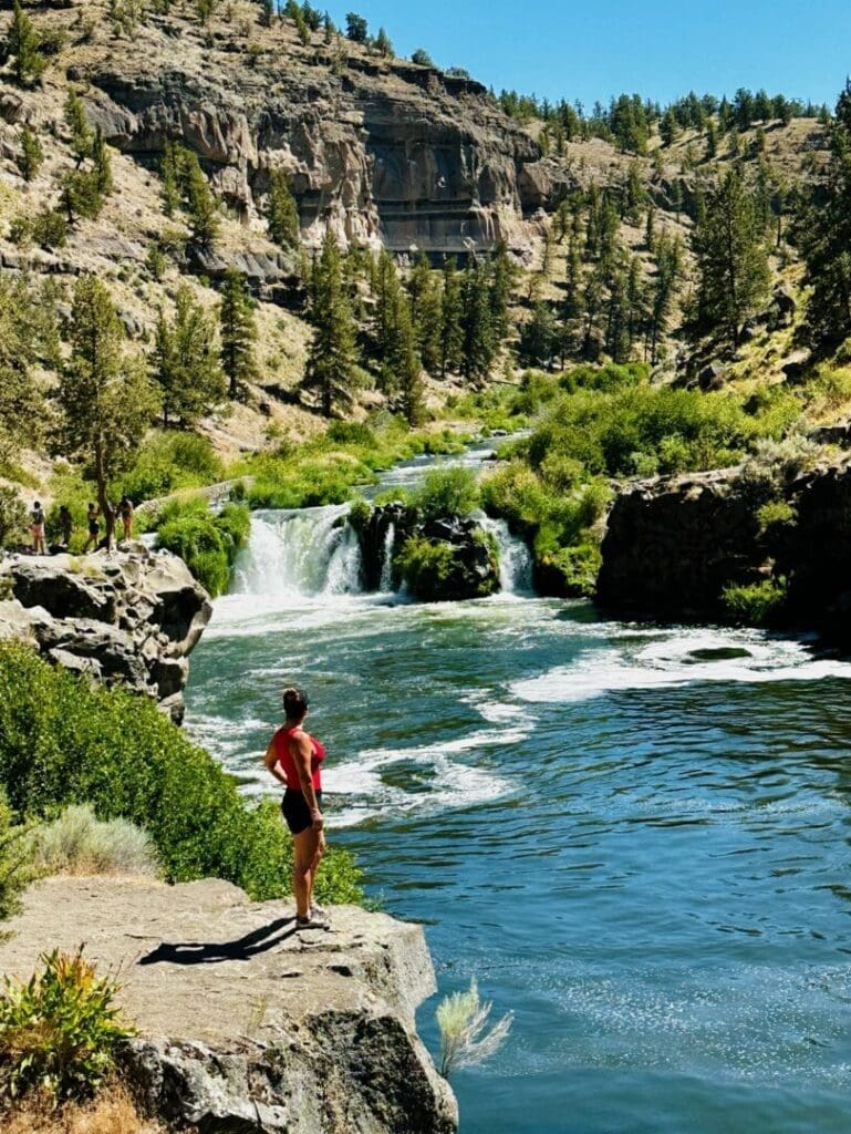 a Person in a Red Shirt and Black Shorts Stands on a Rocky Ledge by Steelhead Falls Gazing at a Small Waterfall Green Trees and Shrubs Surround the Scene with Steep Rocky Hills Under a Clear Blue Sky in the Background