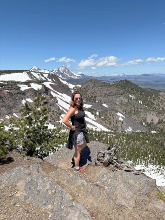 a Woman in Athletic Clothing and Sunglasses Stands Smiling on a Rocky Mountain Ledge Behind Her Are Snowy Peaks Green Forests and a Clear Blue Skycapturing the Adventure of Hiking Near an Airbnb in Bend Oregons Stunning Wilderness