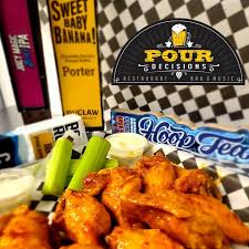 A black basket lined with checkered paper holds saucy buffalo chicken wings, celery sticks, and two cups of ranch. Behind the basket are craft beer cans and a visible Pour Decisions sign—serving up flavor in New Market MD.