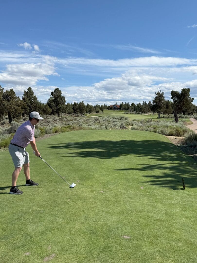 a Person Wearing a Cap Light Shirt and Shorts Stands on a Tee at Pronghorn Golf Course Preparing to Swing a Golf Club the Green Fairway Stretches Ahead Surrounded by Trees and Shrubs Under a Blue Sky with Scattered Clouds