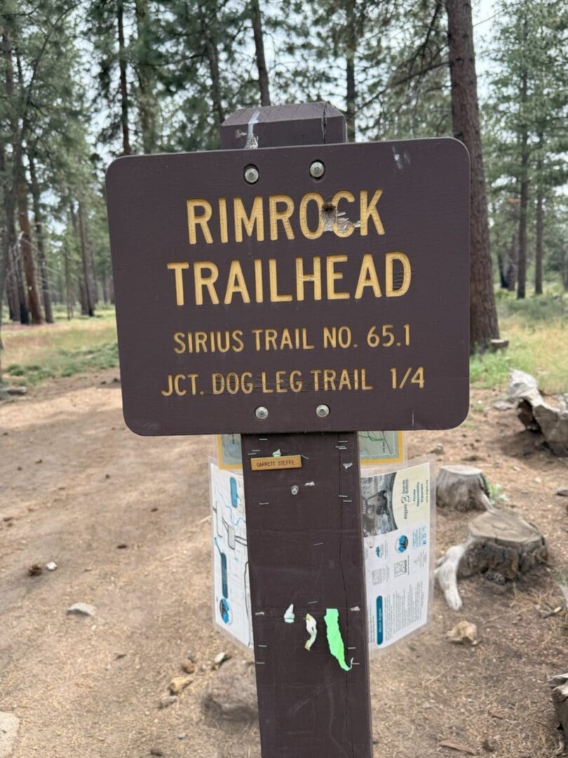 A brown wooden sign reads “Rimrock Trailhead, Sirius Trail No. 65.1, Jct. Dog Leg Trail 1/4.” Surrounded by pine trees and a trail map, this scenic spot is perfect for those seeking classic Bend hikes or memorable hikes near Bend Oregon.