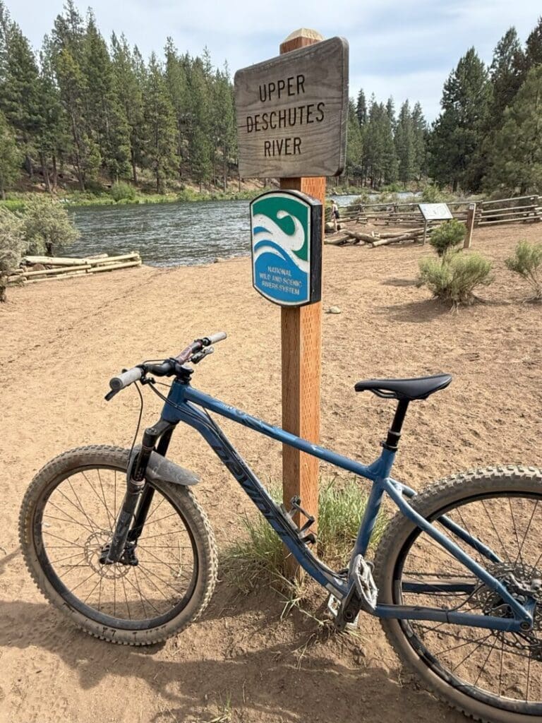 a Blue Mountain Bike is Parked on a Dirt Path by a Wooden Sign Reading Upper Deschutes River Beside the Flowing River with Pine Trees and Fences Nearby the Rimrock Trail Runs Under a Partly Cloudy Sky