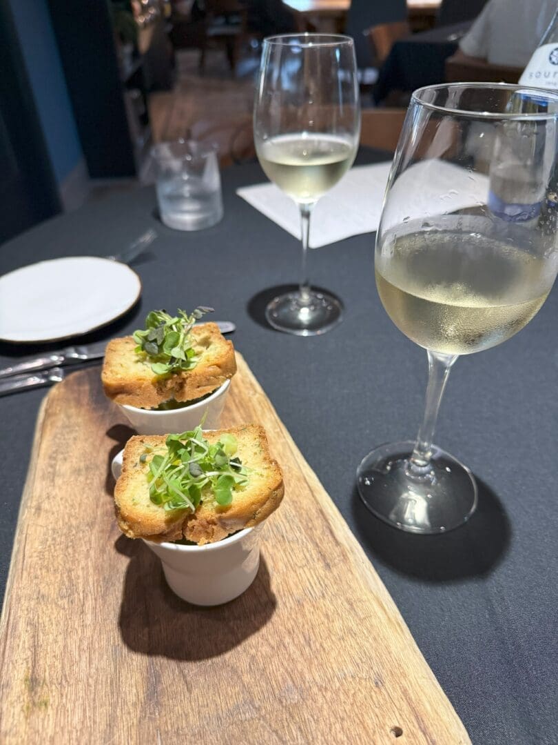 Two Glasses of White Wine Sit on a Black Tablecloth at Market Table Bistro Beside a Wooden Board Holding Two Small White Dishes Each Dish is Topped with Toasted Bread and Fresh Microgreens with a White Plate Cutlery and a Glass of Water in the Background