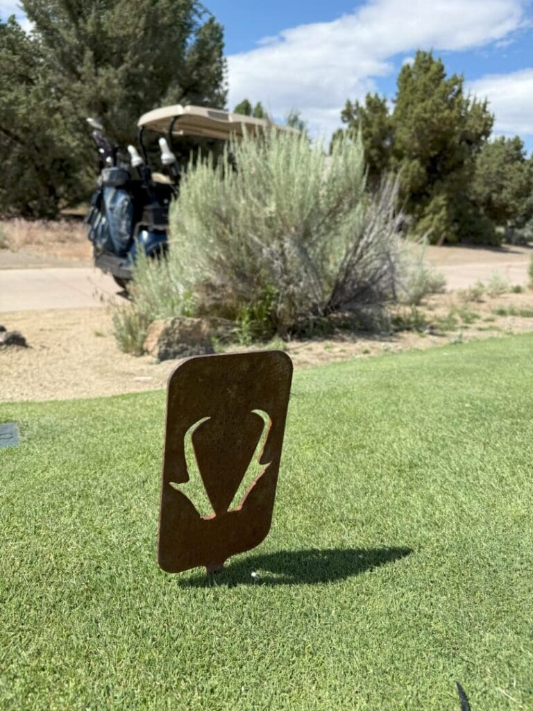 a Metal Tee Marker with an Antelope Horn Design Stands on a Green Pronghorn Golf Course Tee Box in the Background Are Golf Bags on a Cart Bushes Rocks and Trees Beneath a Partly Cloudy Sky Highlighting This Scenic Outdoor Golf Experience