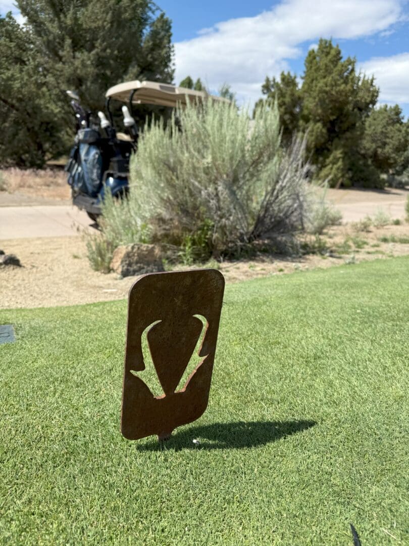 a Metal Tee Marker with an Antelope Horn Design Stands on a Green Pronghorn Golf Course Tee Box in the Background Are Golf Bags on a Cart Bushes Rocks and Trees Beneath a Partly Cloudy Sky Highlighting This Scenic Outdoor Golf Experience
