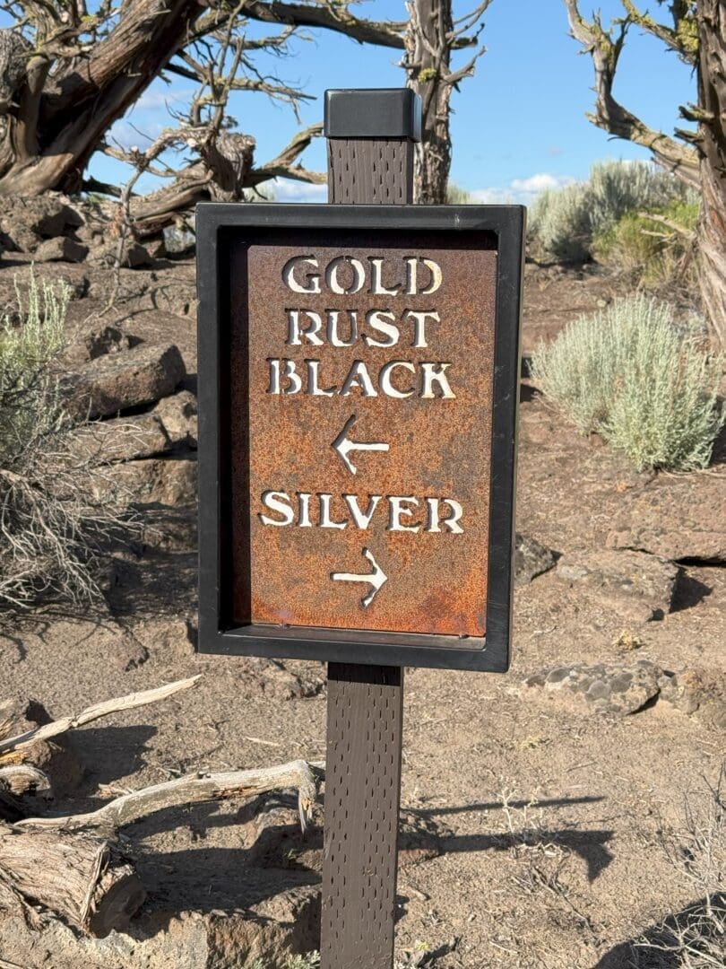 a Weathered Metal Sign on a Post Reads Gold Rust Black with a Left Arrow and Silver with a Right Arrow Set Outdoors Near Pronghorn Golf Course It Stands in a Dry Rocky Area Dotted with Sparse Shrubs and Twisted Tree Branches