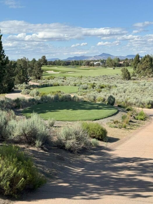 A golf course in Bend, Oregon features a green tee box in the foreground, surrounded by sagebrush and sandy paths. The fairway, trees, and distant mountains are visible under a blue sky with scattered clouds.