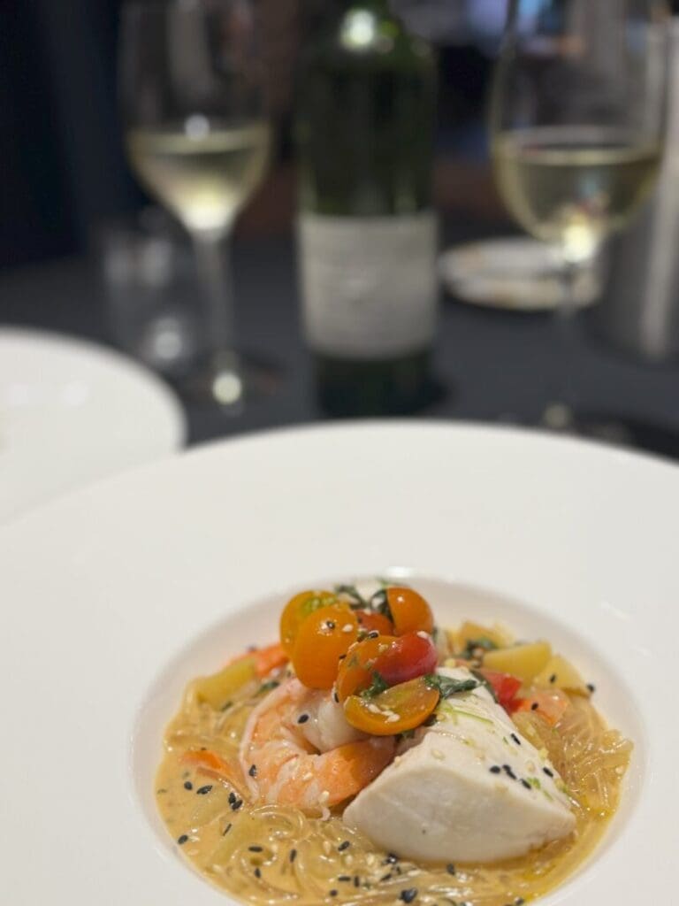 a Close Up of a Plated Gourmet Dish from Market Table Bistro Featuring White Fish Shrimp Colorful Cherry Tomatoes and Sesame Seeds over Noodles in a Creamy Sauce in the Blurred Background Two Glasses of White Wine and a Wine Bottle Are Visible on a Dark Table