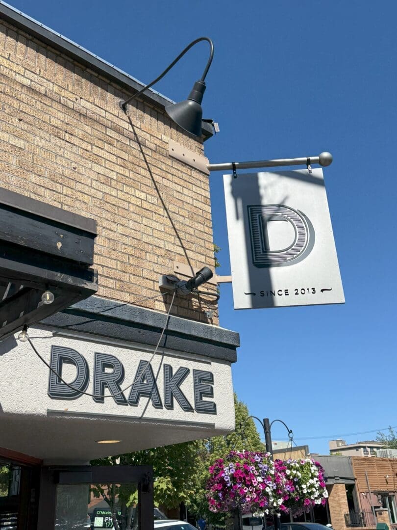 a Beige Brick Building with a Black Sign Reading drake Above the Entrance One of the Standout Bend Restaurants a White Hanging Sign with a Large d and since 2013 is Above It Surrounded by Flowers Street Lamps and a Clear Blue Sky