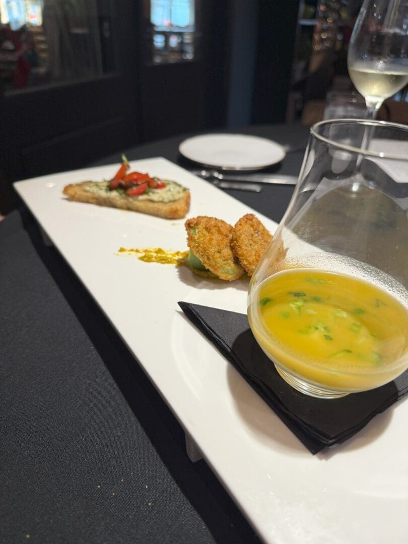at Market Table Bistro a White Rectangular Plate Holds a Glass of Yellow Soup with Herbs Two Breaded Croquettes and a Slice of Toasted Bread Topped with Diced Vegetables the Plate Rests on a Dark Tablecloth Wine Glasses Blurred in the Background
