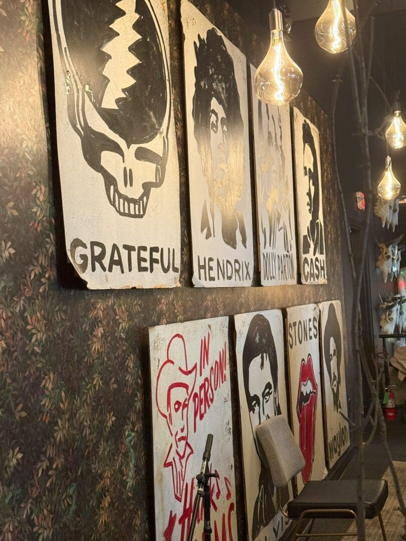 a Wall with Six Large Stylized Black and White Portraits of Famous Musicians Including Grateful Dead Hendrix Dolly Parton Johnny Cash Hank Williams and the Rolling Stones Exposed Bulbs Hang from the Ceiling and a Chair and Mic Stand Sit Below the Paintings