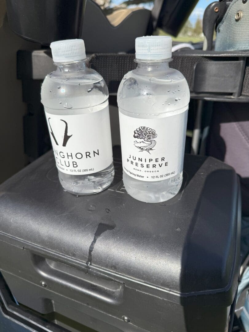 Two Clear Plastic Water Bottles with White Labels Sit on a Black Surface the Left Label Reads Longhorn Club with a Horn Logo Perfect for Pronghorn Golf Course the Right Juniper Preserve with a Juniper Tree Logo Both Are Half Full and Have Condensation