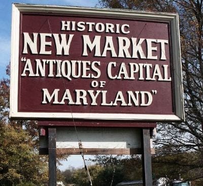 A large maroon and white roadside sign reads: Historic New Market Antiques Capital of Maryland. The sign stands on wooden posts among trees and blue sky, marking the charm of New Market MD as a notable destination.