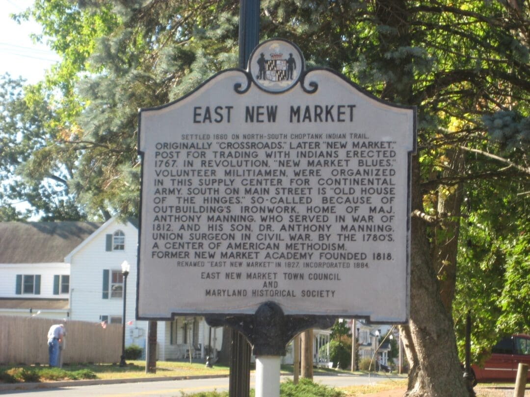 A historical marker for East New Market, MD, stands by a street. The sign details the town’s origins, role in the American Revolution and War of 1812, and mentions Dr. Anthony Manning. Trees and a house are visible in the background on a sunny day.