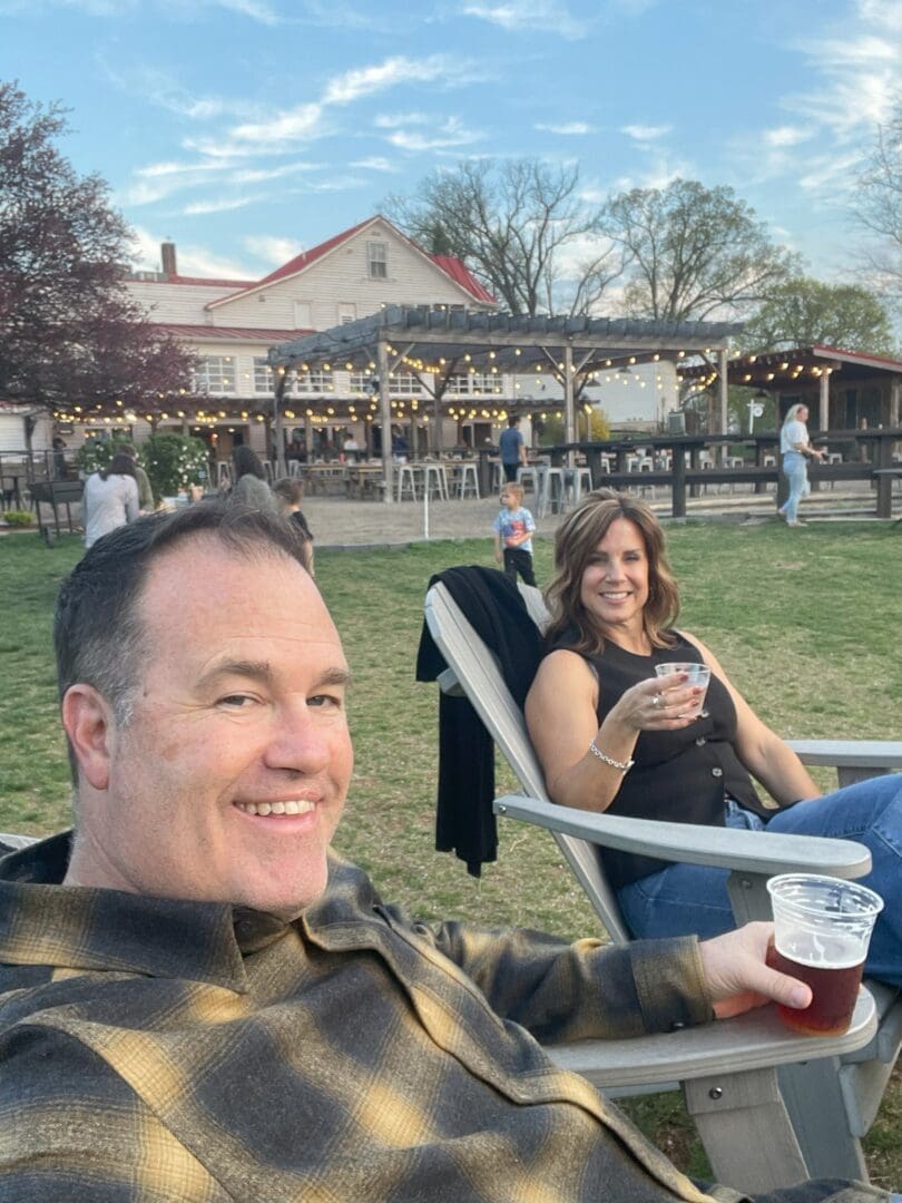A man and woman sit on lawn chairs outdoors, smiling and holding drinks. Behind them is a large farmhouse-style building with string lights, people walking, and trees—all creating a relaxed vibe that invites you to work with us in this cheerful setting.