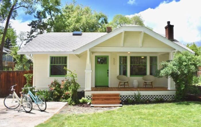 A charming, single-story cream house with a green door and matching window frames in Bend, Oregon. Perfect for an Airbnb stay, it features a cozy porch, two bikes, and is surrounded by a lush lawn under the bright blue sky.