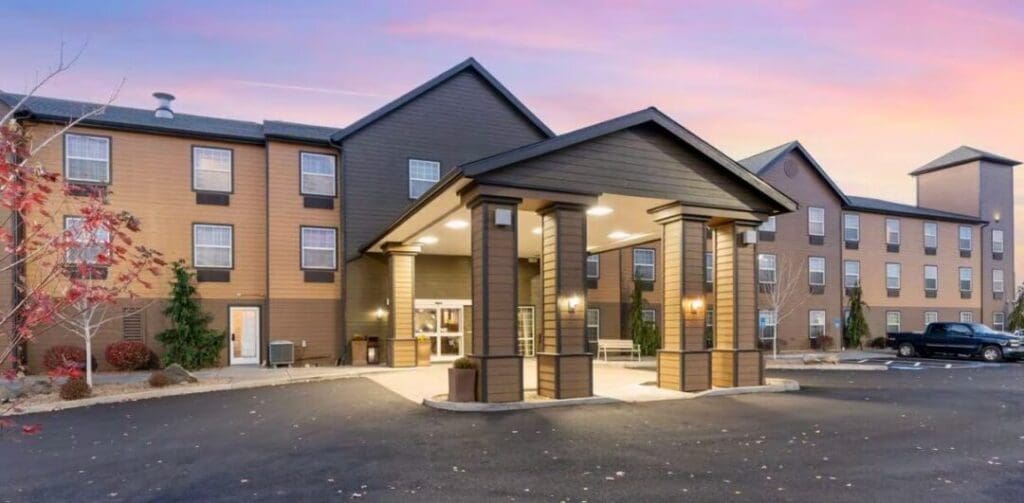a Modern Three Story Hotel with Tan and Brown Siding Stands Under a Pink and Blue Sunset Skyan Inviting Choice for Those Seeking Where to Stay in Bend Oregon the Entrance Features a Covered Driveway and Landscaped Shrubs by the Parking Lot