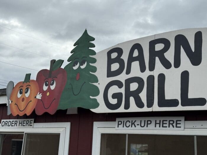 a Sign for barn Grill at Gaver Farm Features Cartoon Faces on a Pumpkin an Apple and a Pine Tree Above Two Windows Labeled Order Here and Pick up Here the Sky is Cloudy and the Red Building Has White Trim