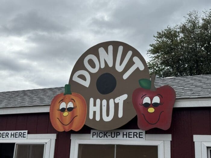 a Round Sign Reading Donut Hut with a Donut Hole in the Center is Displayed at Gaver Farm Beside the Sign Are Cartoon Faces of a Smiling Pumpkin and Apple the Sky is Cloudy with order Here and pick up Here Signs Below