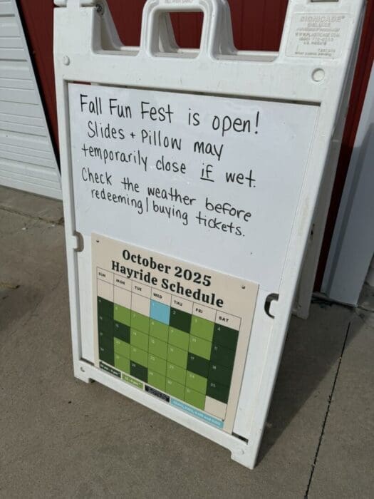 a White Sandwich Board Sign Stands on the Gaver Farm Sidewalk the Top Has Handwritten Text About Fall Fun Fest Noting Slides and Pillow May Close if Wet Below is an October 2025 Hayride Schedule Calendar in Green Shades