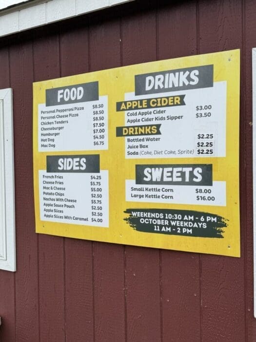 a Yellow and White Menu Board on a Red Wooden Wall at Gaver Farm Displays Prices for Food Drinks Sides and Sweets Items Include Pizza Chicken Tenders Cheeseburgers Fries Apple Cider Popcorn and More Plus Weekend and Weekday Hours