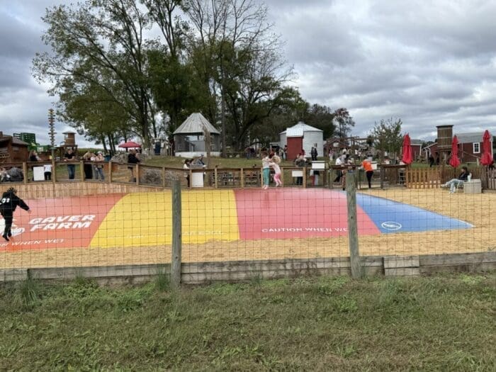 a Large Colorful Jumping Pillow with Red Yellow and Blue Sections Sits in a Sandy Area at Gaver Farm Surrounded by a Wooden Fence Children Play on the Pillow As Adults Watch Nearby Among Trees Picnic Tables Farm Buildings and Cloudy Skies