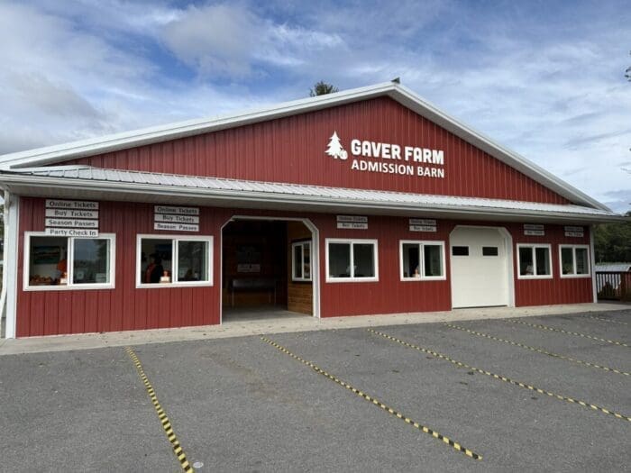 A red barn-style building with white trim labeled “Gaver Farm Admission Barn” stands under a partly cloudy sky in Mt Airy, MD. Signs above the windows mention buying tickets and season passes, while yellow lines mark parking spaces in the foreground.