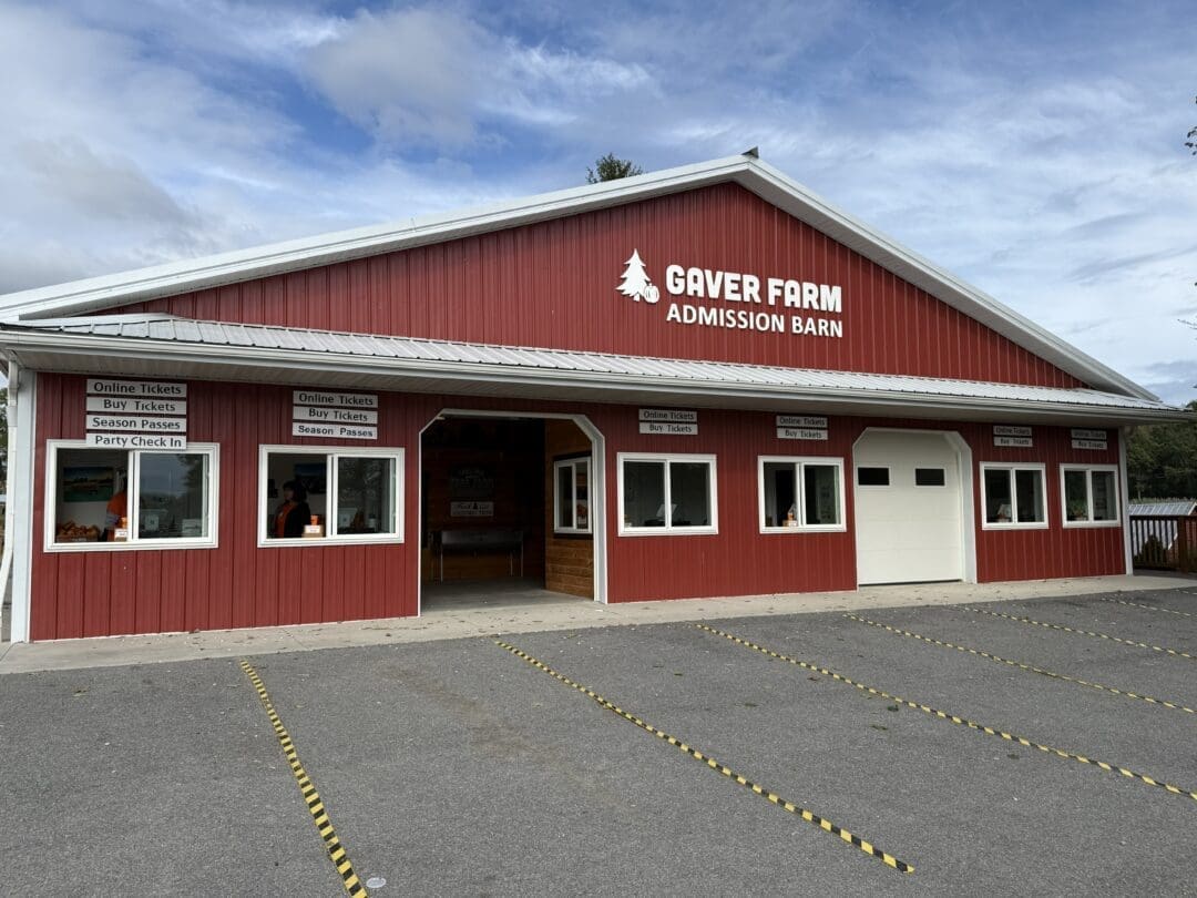 A red barn-style building with white trim labeled “Gaver Farm Admission Barn” stands under a partly cloudy sky in Mt Airy, MD. Signs above the windows mention buying tickets and season passes, while yellow lines mark parking spaces in the foreground.