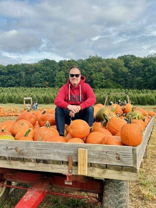 A man wearing sunglasses, a red hoodie, jeans, and sneakers sits cross-legged on a wooden wagon filled with orange pumpkins in a pumpkin patch in Frederick, MD—one of the fun things to do in Frederick on a cloudy day.