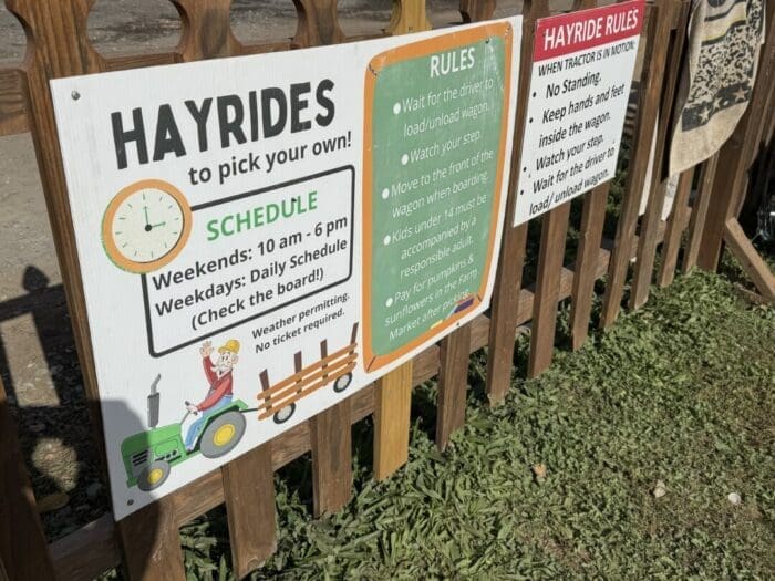 a Wooden Fence at Gaver Farm Displays Three Signs About Hayrides the Largest Details Gaver Farm Hayride Schedules with a Drawing of a Tractor Pulling Hay Bales the Others List Rider Instructions and Safety Precautions with Grass Below the Signs