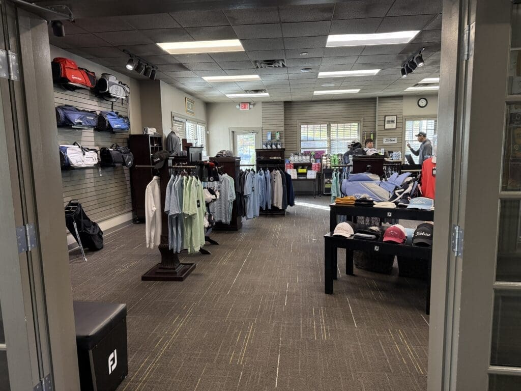 a Golf Pro Shop Interior at the Links at Challedon with Racks of Clothing Hats and Golf Bags on Display Two People Stand at the Checkout Counter Near the Back the Shop Features Carpeted Floors Bright Lighting and Large Windows Throughout