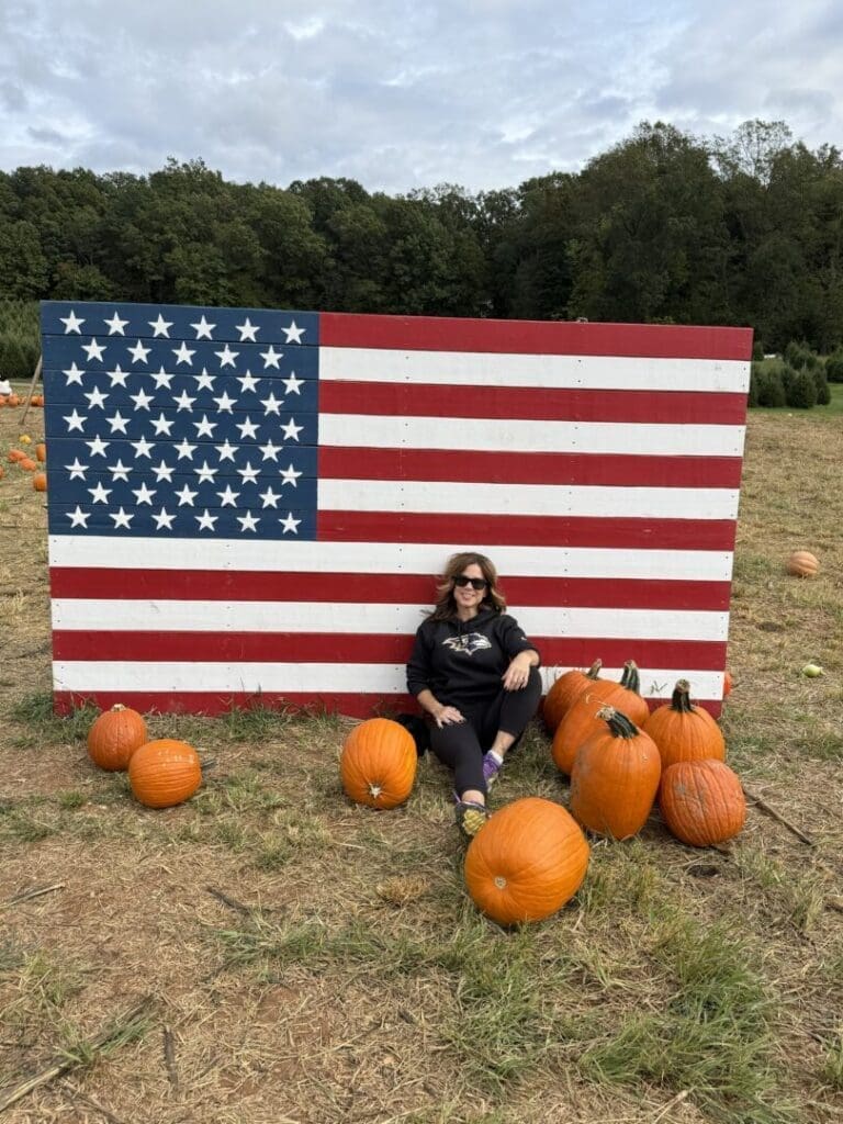 A woman wearing sunglasses sits on the ground at Gaver Farm, leaning against a large wooden American flag display. She is surrounded by several pumpkins on dry grass in a pumpkin patch, with trees and a cloudy sky in the background.