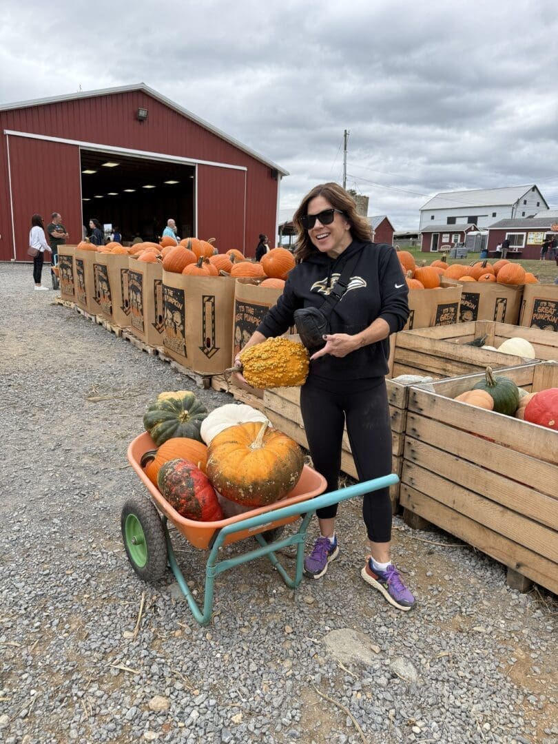 a Woman in Sunglasses and Athletic Wear Smiles While Holding a Large Yellow Gourd Beside a Wheelbarrow Full of Pumpkins at Gaver Farms Outdoor Pumpkin Patch Behind Her Are Crates of Pumpkins and a Red Barn Under a Cloudy Sky