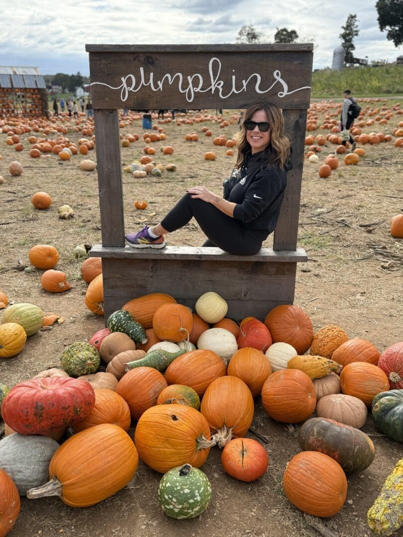 a Woman with Wavy Brown Hair Wearing Sunglasses a Black Outfit and Purple Shoes Sits Smiling in a Wooden Frame Labeled Pumpkins at Gaver Farm She is Surrounded by Orange and Multicolored Pumpkins at a Lively Pumpkin Patch