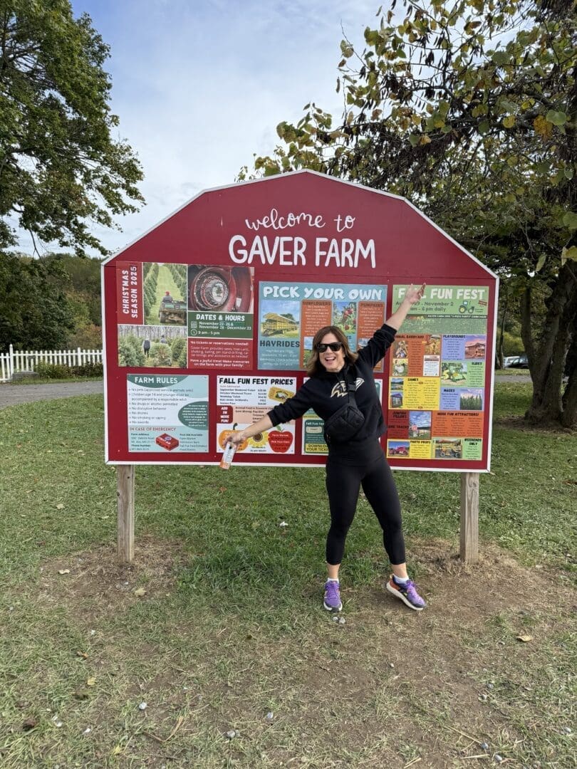 a Woman in Sunglasses Black Clothes and Purple Sneakers Stands Smiling and Pointing at a Large Red Welcome to Gaver Farm Sign Filled with Colorful Information and Farm Rules She Holds a Gaver Farm Flier Standing on Grass Near Trees and a White Fence