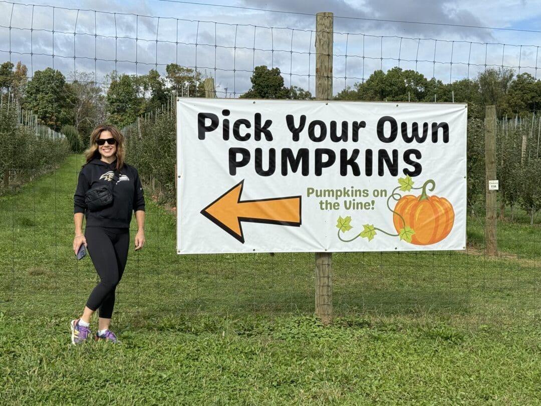 a Woman in Sunglasses and Athletic Wear Stands Smiling Beside a Large Pick Your Own Pumpkins Sign at Gaver Farm on a Grassy Field Pumpkin and Vine Illustrations Decorate the Sign Trees Line the Background Under a Cloudy Sky