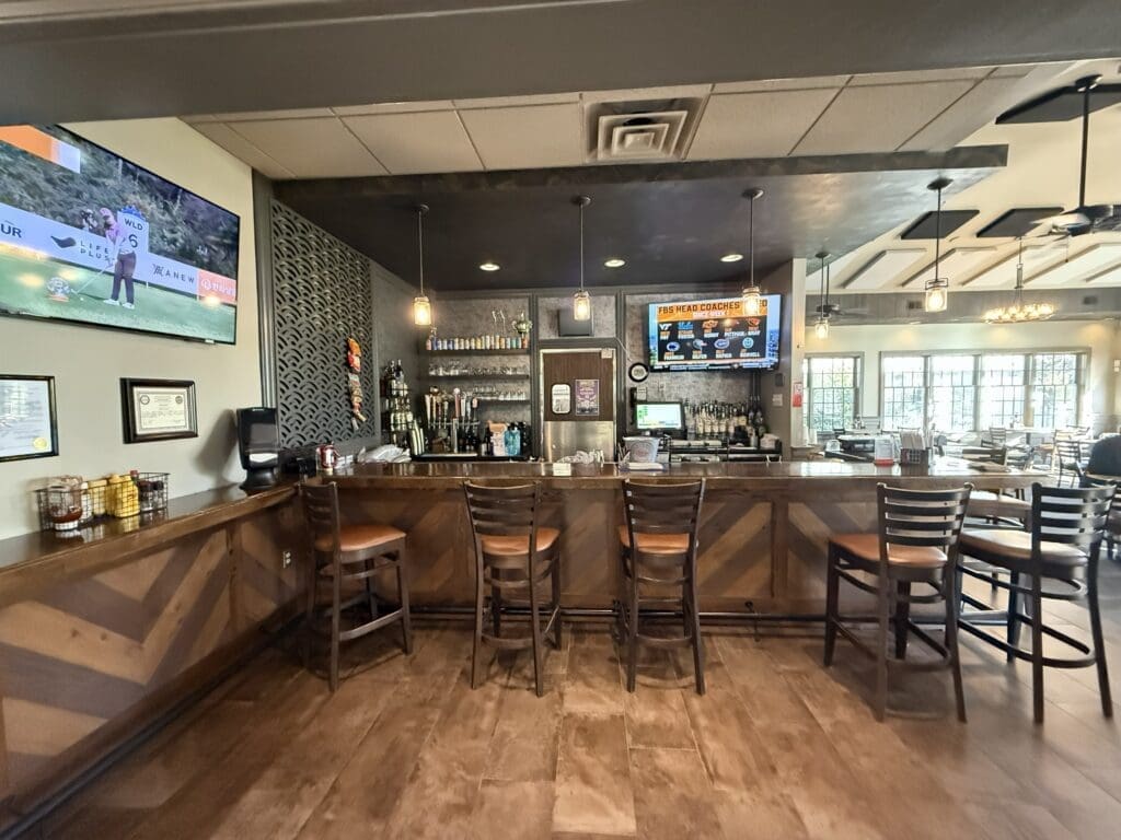a Modern Bar at the Links at Challedon Features a Wooden Counter and Four High Chairs Shelves with Bottles and Glasses Line the Back Two Digital Menu Screens Glow and a Tv on the Left Wall Shows Golf Large Windows on the Right Fill It with Light
