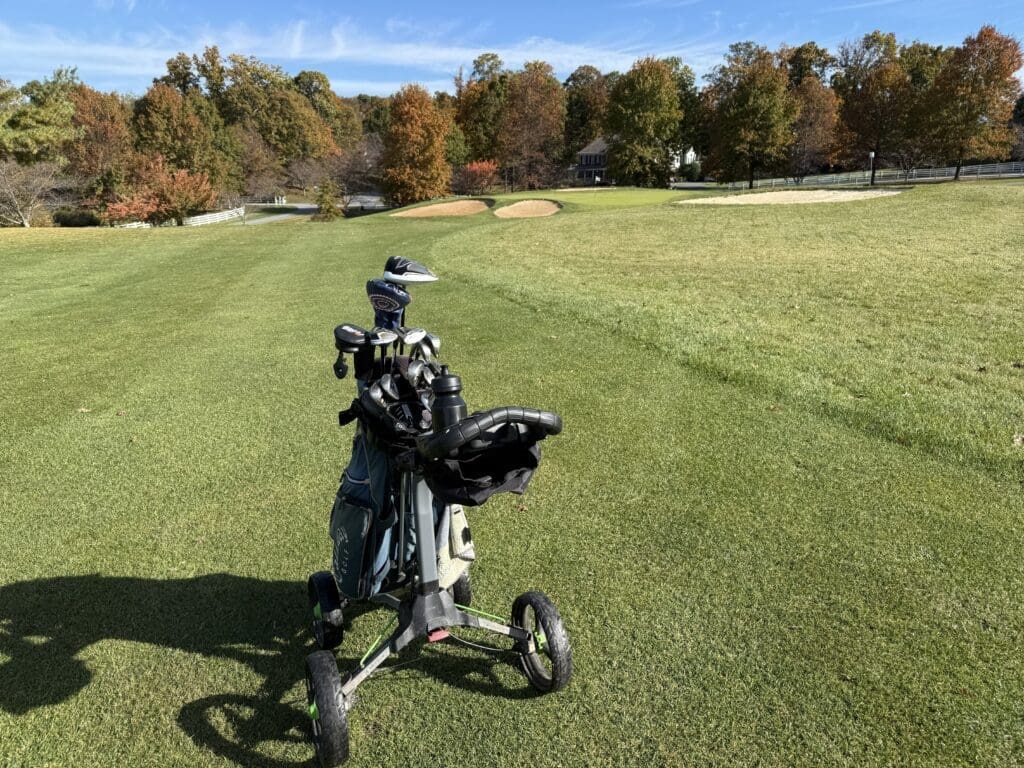 a Golf Pushcart Holding Several Clubs Sits on a Lush Green Fairway at the Links at Challedon in the Distance Sand Bunkers Autumn Colored Trees and a House Appear Under a Blue Sky with Wispy Clouds and Gentle Morning Shadows