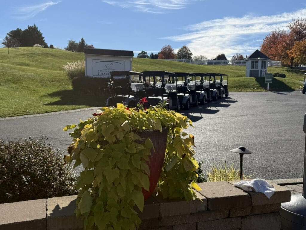 a Row of Golf Carts is Parked on a Paved Lot at the Links at Challedon Near a Grassy Hill with a White Sign and Small Building a Large Planter with Green and Yellow Plants Sits in the Foreground Beneath Blue Wispy Skies and Autumn Trees