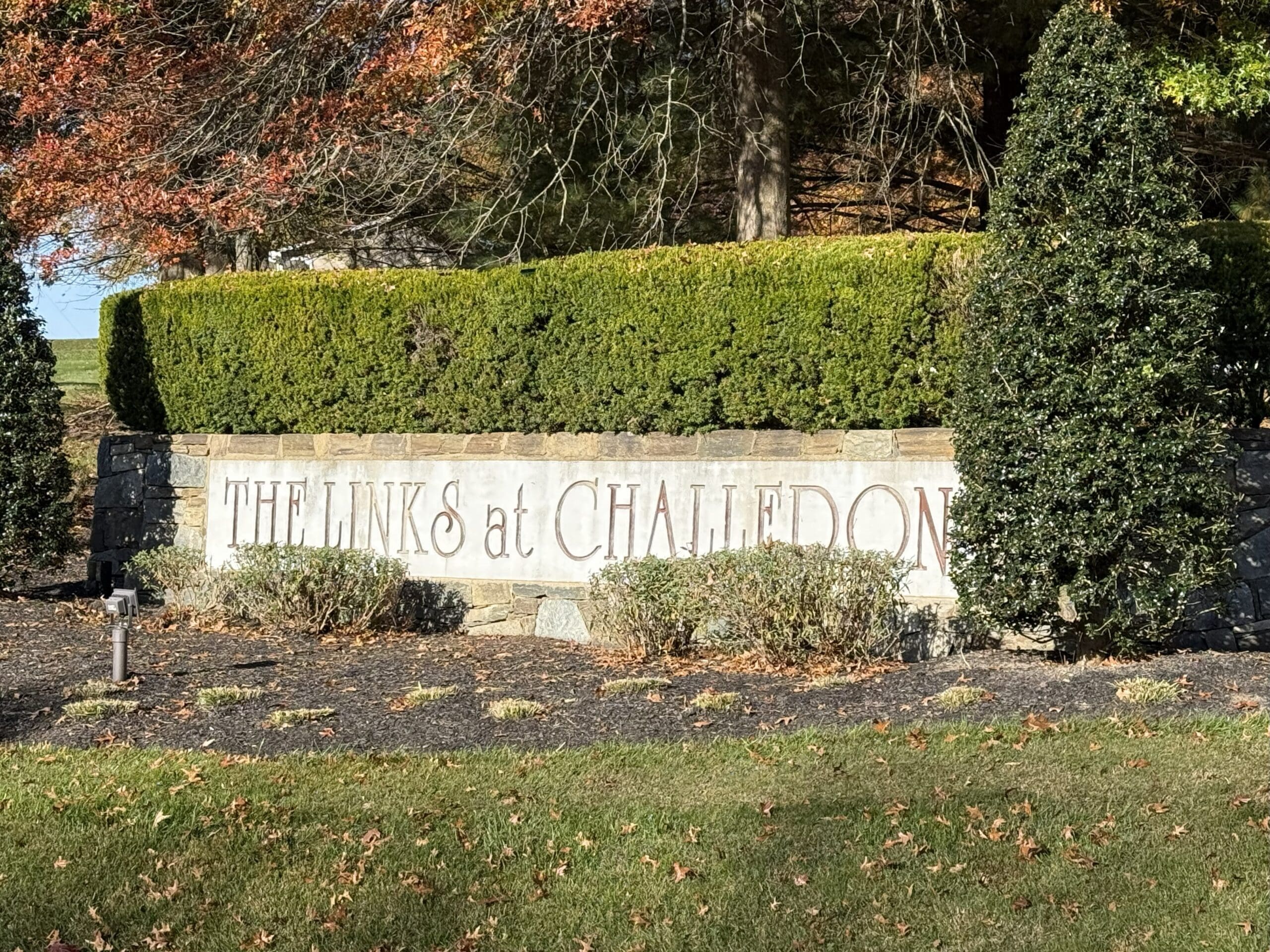 a Stone Sign with Neatly Trimmed Hedges and Two Tall Bushes Displays the Words the Links at Challedon Autumn Leaves and Sunlight Add Warmth to the Scene While a Small Light Fixture Highlights the Links at Challedons Welcoming Entrance