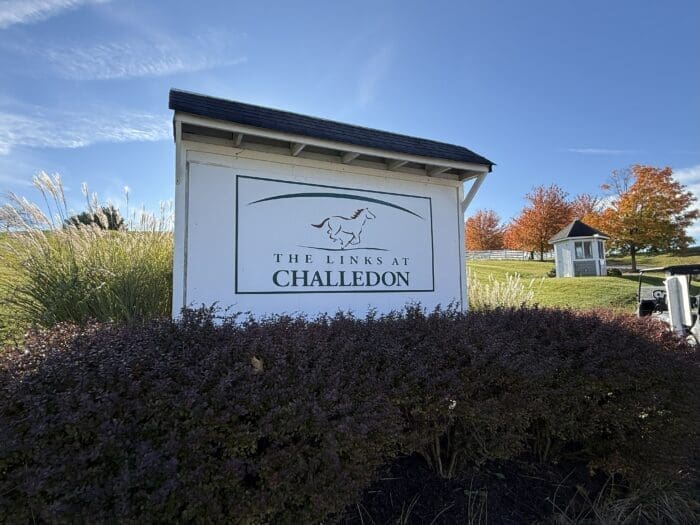A white sign with a horse logo and text reading “The Links at Challedon” stands among shrubs and tall grass in Mt Airy MD. In the background, autumn trees with orange leaves and a small shed sit under a bright blue sky with wispy clouds.