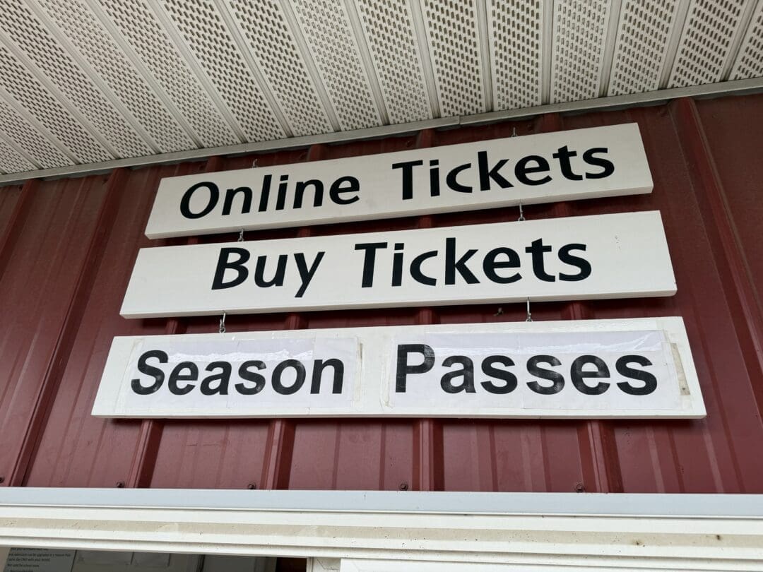 Three White Rectangular Signs with Black Text Hang on a Red Metal Wall Under a Roof at Gaver Farm the Top Sign Reads Online Tickets the Middle Says Buy Tickets and the Bottom Reads Season Passes Guiding Visitors to Ticketing Options