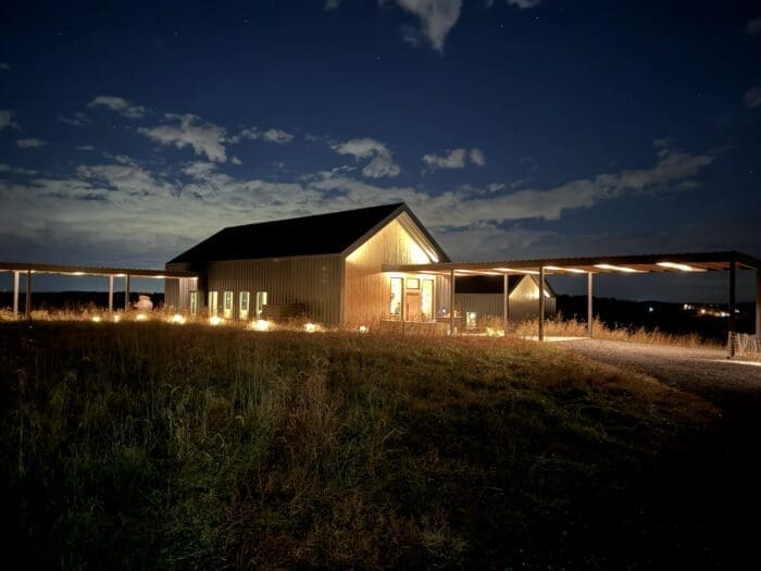 A warmly lit barn-style building stands alone at night under a partly cloudy sky in Mt Airy MD. Soft lights illuminate windows and a covered walkway, while tall grass surrounds the structure, creating a peaceful rural scene.