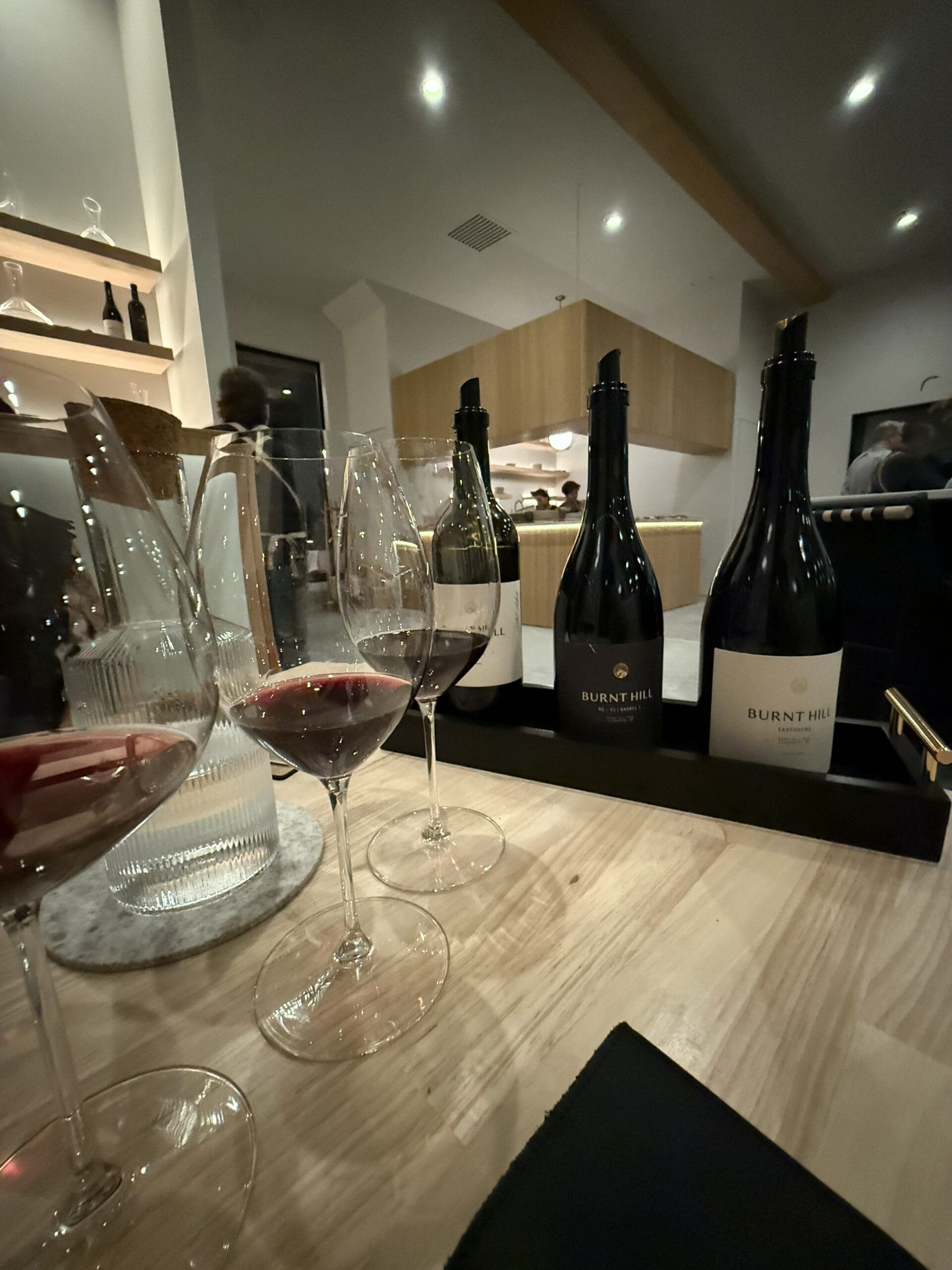 a Wooden Table with Three Glasses of Red Wine a Glass Water Carafe and Three Burnt Hill Farm Wine Bottles the Background Features a Modern Softly Lit Interior with Shelves People and a Wooden Ceiling Beam