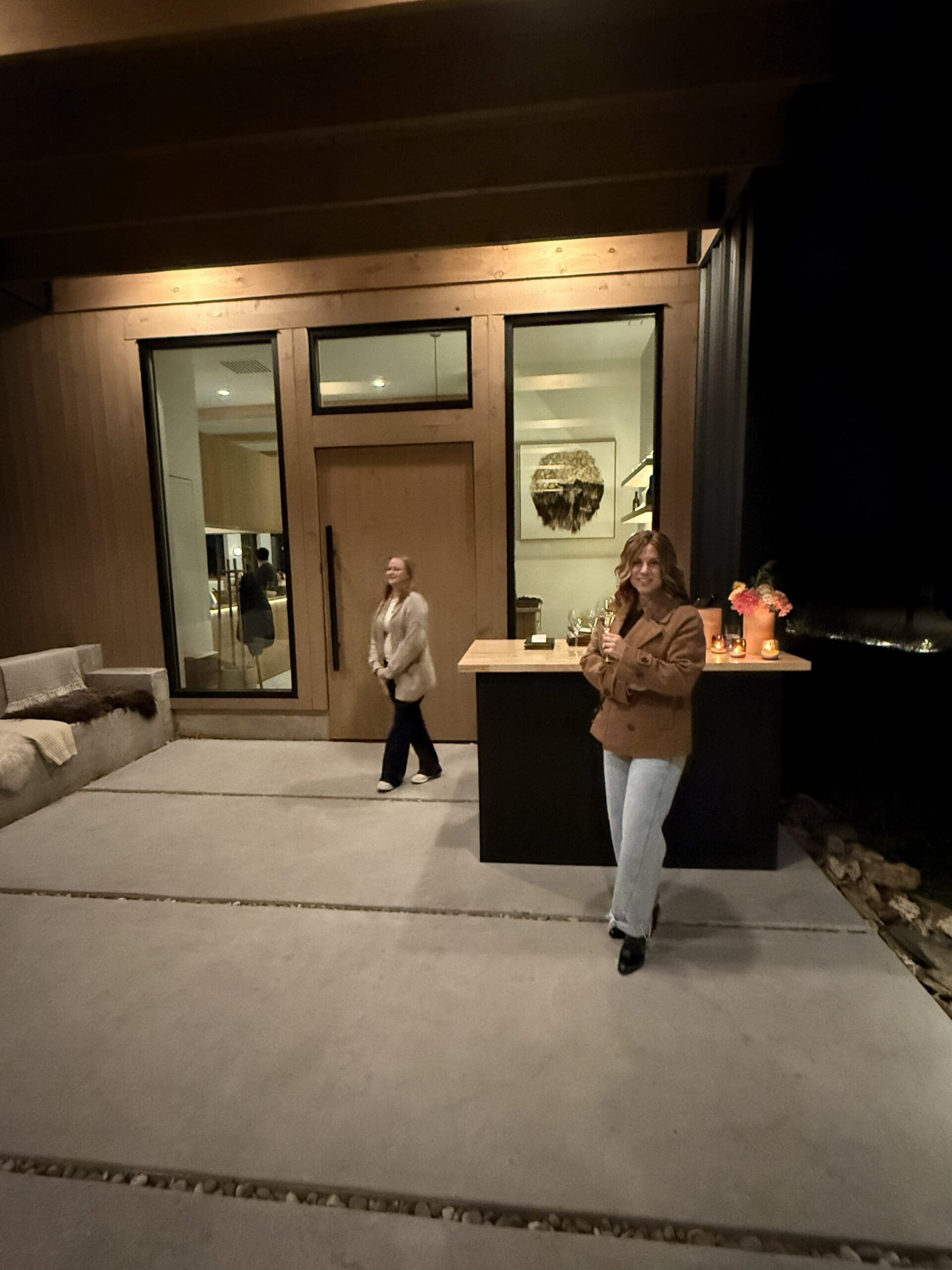 Two Women Stand Outside Burnt Hill Farm at Night One Holds a Drink Near a Small Table with Flowers and Bottles the Other Stands Closer to the Door Warm Lighting Highlights the Modern Houses Large Windows and Wooden Paneling